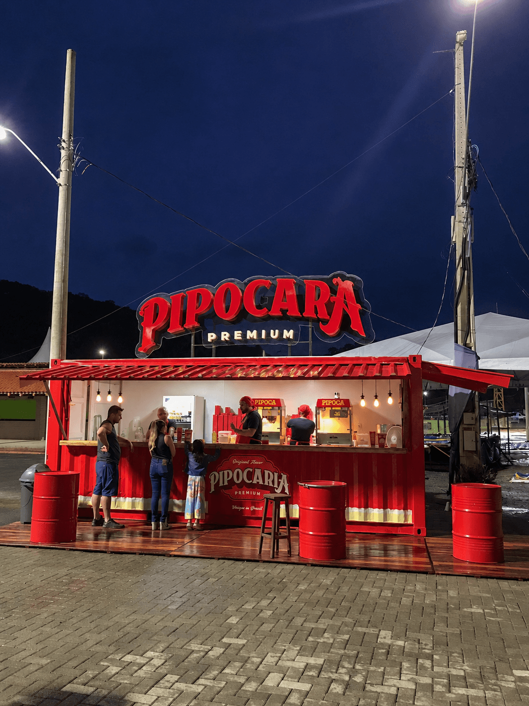 Container Pipocaria Premium 20 pés à noite com letreiro neon PIPOCARIA PREMIUM iluminado e atendentes